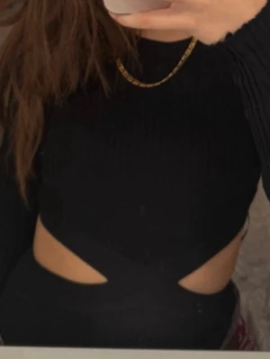 Svart croppad bodymed cut-outs - Säljer en svart croppad topp med långa ärmar och trendiga cut-outs vid midjan. Toppen har en tight passform och är ribbad för extra stilpoäng. Perfekt för dig som vill sticka ut lite extra.