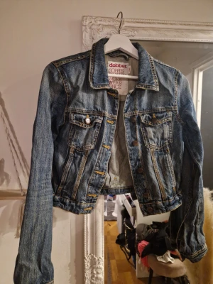 Klassisk blå jeansjacka från Dobber - Snygg blå jeansjacka från Dobber i klassisk modell med två bröstfickor och knappar framtill. Jackan har kontrastsömmar och en cool, lite croppad passform. Perfekt att slänga över en hoodie eller t-shirt för en avslappnad look. 