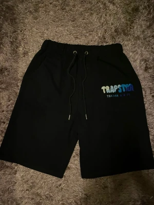Trapstar shorts - Svarta shorts från Trapstar med elastisk midja och snörning. Tryck i vitt och blått på vänster ben. Sköna och avslappnade, perfekta för sommaren. Tillverkade i mjukt material som känns bekvämt hela dagen.