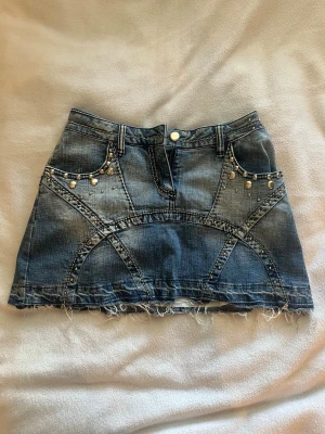Kort jeanskjol med nitar och rå kant - Säljer en blå kort jeanskjol med coola nitar och dekorativa sömmar. Kjolen har rå, fransig kant nertill och fickor fram. Perfekt för dig som gillar en edgy och trendig stil. 