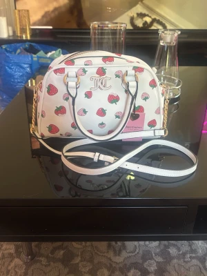 Vit handväska med jordgubbsmönster - Söt vit handväska från Juicy Couture med färgglatt jordgubbsmönster och guldfärgade detaljer. Väskan har både handtag och en justerbar axelrem i vitt, samt en JC-logga framtill. Har aldrig använt den du kan se prislappen . Köpte för 99 dollar men säljer för 800kr