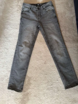 Gråa slim fit jeans Jack & Jones - Snygga gråa slim fit jeans från Jack & Jones med klassisk femficksdesign och diskreta slitningar. Jeansen har raka ben och är tillverkade i ett mjukt denimtyg som ger en skön passform. Perfekta för en avslappnad och trendig look.