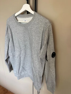 Grå sweatshirt från C.P. Company - Snygg grå sweatshirt från C.P. Company med rund halsringning och ribbade muddar. Tröjan har en ikonisk svart linsdetalj på ärmen och är tillverkad i mjukt bomullsmaterial. Perfekt för en avslappnad och trendig look..                                Använd någon gång endast! Har ej kvitto!  