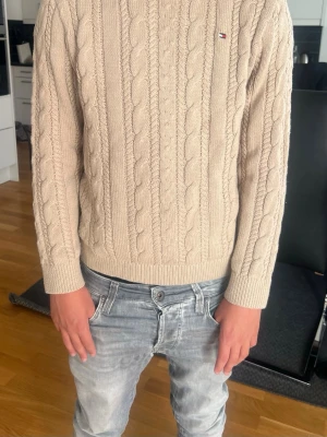 Beige kabelstickad tröja Tommy Hilfiger - Snygg beige kabelstickad tröja från Tommy Hilfiger med rund halsringning och diskret logga på bröstet. Tröjan har långa ärmar och ribbade muddar vid ärmslut och nederkant. Perfekt för dig som gillar klassisk och stilren look.