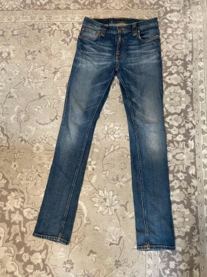 Blå jeans från Nudie Jeans co - Säljer ett par klassiska blå jeans från Nudie Jeans med raka ben och snygga slitningar. Jeansen har orange kontrastsömmar, fem fickor och knappgylf. Perfekta för dig som gillar en tidlös och avslappnad stil.