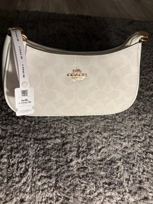  Coach väska - Stilren vit handväska från Coach med guldfärgade detaljer och präglad logotyp framtill. Väskan har ett diskret mönster med Coachs signatur-C och en kort axelrem i vitt läder med guldfärgade kedjor. Perfekt för dig som gillar klassisk och trendig design.