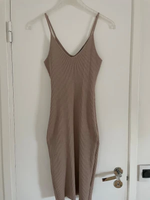 Stickad klänning - Säljer en beige ribbad långklänning med smala axelband och v-ringning. Klänningen har en enkel och stilren design i stretchigt material som sitter snyggt!! Köpt på marknad utomlands!!