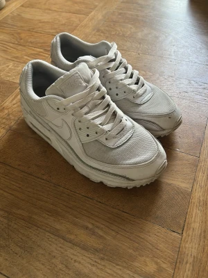 Vita Nike Air Max 90 sneakers - Säljer ett par klassiska vita Nike Air Max 90 sneakers med mesh och läderdetaljer. Skorna har en chunky sula och ikonisk swoosh på sidan. Perfekta för dig som gillar streetwear och sportig stil.