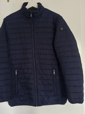 Mörkblå pufferjacka från True North - Säljer en mörkblå pufferjacka från True North Street Wear. Jackan har två dragkedjeförsedda fickor, hög krage och quiltad design. Perfekt för kalla dagar och har en stilren look med diskret logga på ärmen.