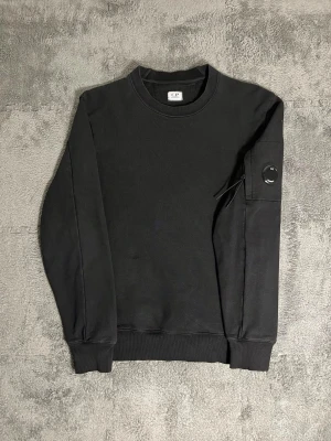 C.P. Company – Black Lens Sweatshirt (M) - Sleek and modern C.P. Company sweatshirt in classic black, featuring the brand’s signature lens detail on the sleeve — the same iconic design as the white version. Size M, length 66 cm, pit‑to‑pit 53 cm – comfortable and structured fit.  ✅ Condition: Excellent – no visible wear or stains 💯 Authenticity: 100 % genuine, can be verified on certilogo.com   📦 Shipping: Fast dispatch after purchase  This sweatshirt captures C.P. Company’s technical streetwear aesthetic — minimalist, durable.
