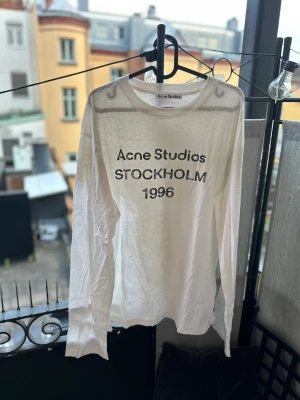 Vit långärmad tröja Acne Studios - Vit långärmad topp från Acne Studios med svart text 'Acne Studios STOCKHOLM 1996' på bröstet. Rund halsringning och rak passform. Perfekt för dig som gillar minimalistisk och stilren design.