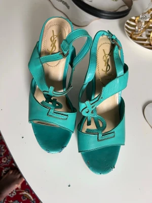Turkosa sandaler från Yves Saint Laurent - Säljer ett par snygga turkosa sandaler från Yves Saint Laurent med öppen tå och justerbart spänne vid ankeln. Sandalerna har YSL-loggan framtill och är tillverkade i läder med en cool retrovibe. Perfekta för dig som vill sticka ut med färg och stil.