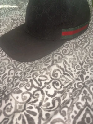 Keps - Snygg svart keps med diskret mönster och en grön-röd rand på sidan. Klassisk böjd skärm och justerbar passform. Perfekt accessoar för en trendig streetwear-look.