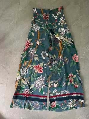 Blommiga byxor i viskos GP&J Baker x H&M - Supersnygga byxor med vida ben från GP&J Baker x H&M. Mörkgrön botten med färgglatt blommönster, fåglar och trädgrenar. Dekorativa ränder längst ner på benen. Tillverkade i viskos som känns skönt mot huden.