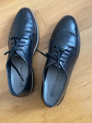 Svarta loafers i skinn från Ecco 40 - Snygga svarta loafers från Ecco i äkta skinn med robust sula och klassisk snörning. Skorna har en stilren design med kontrastsöm längs sulan och en bekväm innersula. Perfekta för dig som vill ha både stil och komfort.