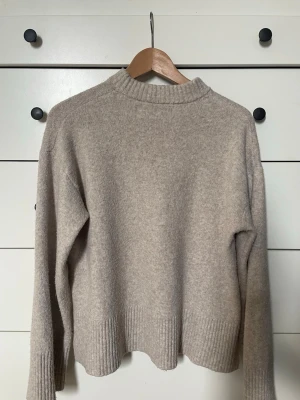 Beige stickad tröja med ribbade detaljer - Mysig beige stickad tröja med hög hals och ribbade muddar vid ärmslut och nederkant. Tröjan har en avslappnad passform och små slitsar i sidorna, vilket ger en modern look. Perfekt för kyliga dagar och enkel att matcha med olika outfits. Lite nopprig men går att fixa 💕