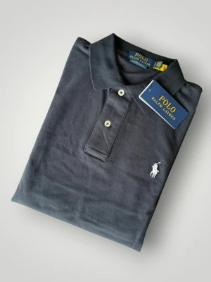 Mörkblå pikétröja från Polo Ralph Lauren - Klassisk mörkblå pikétröja från Polo Ralph Lauren med vit broderad logga på bröstet. Tröjan har krage, korta ärmar och knäppning med två knappar framtill. Perfekt för en stilren och avslappnad look.