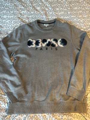 Grå Kenzo Paris sweatshirt - Säljer en grå sweatshirt från Kenzo med broderad logga i blått, vitt och svart på bröstet samt texten PARIS under. Tröjan har rund halsringning, ribbade muddar och är i mjukt material. Perfekt för dig som gillar streetwear och vill ha något stilrent men ändå unikt.