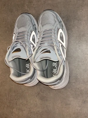 Zara - Säljer ett par grå sneakers från Zara  med reflekterande detaljer och tydligt CD-logo på sidan. Skorna har mesh- och syntetmaterial, tjock vit sula och klassisk snörning. Perfekta för dig som gillar stilrena och trendiga sneakers.
