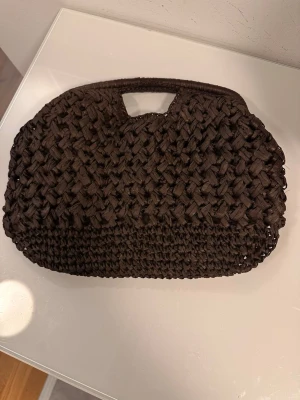 Brun flätad kuvertväska med handtag - Säljer en brun kuvertväska i flätad design med inbyggt handtag. Väskan har en mjuk, rundad form och är tillverkad i ett grovt, textilliknande material. Perfekt för dig som gillar en bohemisk och trendig stil.