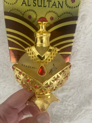 Al Sultan parfym från Khadlaj - Lyxig parfymflaska i guld med röda detaljer och orientaliskt mönster. Flaskan har en rund form och är dekorerad med röda stenar och guldfärgade ornament. Perfekt för dig som gillar exklusiva och unika dofter.
