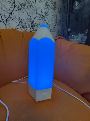Bordslampa formad som penna - Säljer en unik bordslampa i form av en stor penna. Lampan har LED-belysning som kan skifta färg mellan blått och lila. Basen är vit och lampan har en sladd för ström. Perfekt som dekoration till skrivbordet eller rummet.