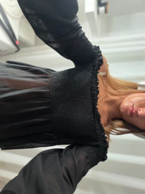 Svart offshoulder blus med smock - Säljer en svart offshoulder blus med smock över bysten och ärmarna. Blusen har långa, genomskinliga ärmar och volangkant upptill. Perfekt för dig som gillar trendiga och feminina toppar. Aldrig använd 