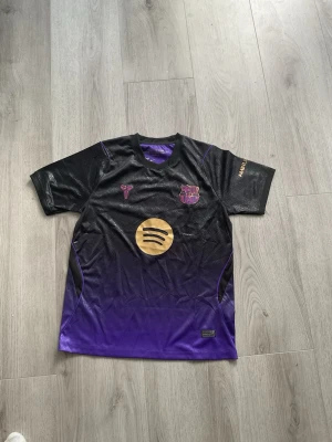 Svart och lila FC Barcelona t-shirt - Snygg FC Barcelona t-shirt i svart och lila med guldfärgad Spotify-logga på bröstet. Tröjan har klubbmärke och Nike-logga framtill samt texten 'AMBILU' på ärmen. Kortärmad modell i syntetmaterial, perfekt för fotboll eller streetwear.