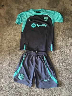 Barcelona träningsset Nike turkos/svart - Säljer ett FC Barcelona träningsset från Nike i svart och turkos. Setet består av en kortärmad tröja med klubbmärke, Nike-logga och Spotify-tryck samt matchande shorts med klubbmärke och turkosa detaljer. Perfekt för fotbollsträning eller gymmet.