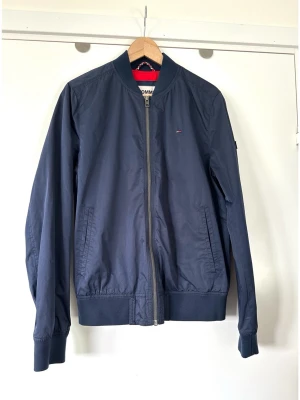 Mörkblå bomberjacka Tommy Hilfiger - storlek S - Stilren marinblå / mörkblå / navy bomberjacka från Tommy Jeans i storlek S. Tidlös modell med Helen dragkedja framtill, ribbade muddar i ärmslut och nederkant samt två praktiska sidofickor. Tommy Hilfiger-loggan finns diskret broderad på bröstet och som en flagga på vänster ärm. En tidlös klassiker som kommer hålla säsong efter säsong. Nypris 2999:-. Mycket gott skick, som ny!
