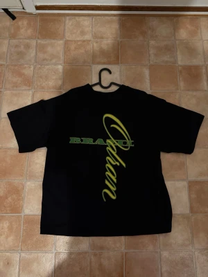 Svart t-shirt med grön text - Säljer en svart t-shirt med coolt tryck på ryggen där det står 'BRAND' och 'Odan' i grön och gul text. På framsidan finns en liten 'Odan'-logga i samma färg. Klassisk passform och rund hals, perfekt till jeans eller shorts.