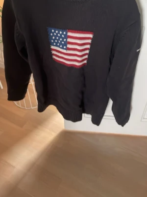 Mörkblå stickad Gant tröja USA-flagga - Mörkblå stickad tröja från Gant med stor amerikansk flagga på bröstet och broderad text 'GANT NEW HAVEN 1949' på ärmen. Tröjan har ribbade muddar och klassisk passform. Perfekt för dig som gillar preppy stil och vill sticka ut.