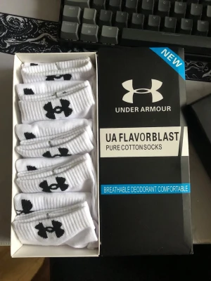 Vita Under Armour bomullsstrumpor - Säljer ett flerpack vita låga strumpor från Under Armour med svart logga på sidan. Strumporna är tillverkade i bomull och har en ribbad kant upptill för extra komfort. Perfekta till sneakers och vardagsbruk.