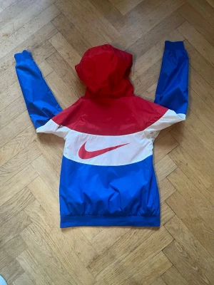 Nike vindjacka - klassisk Nike Sportswear Windrunner vindjacka i färgblock (röd, vit och kungsblå). Perfekt som överdragsjacka till våren, hösten eller svalare sommardagar!Märke: NikeStorlek: M för barn (passar ca 137–147 cm / ca 10–12 år)