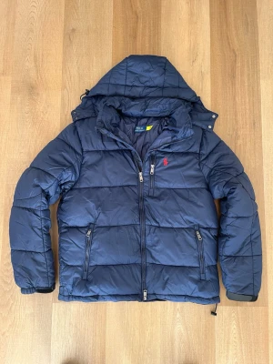 Mörkblå dunjacka från Polo Ralph Lauren - Snygg mörkblå dunjacka från Polo Ralph Lauren med huva och dragkedjor. Jackan har en röd broderad logga på bröstet, två sidofickor med dragkedja och en bröstficka. Perfekt för kalla vinterdagar och riktigt skön att bära. Avklippt storleks tag