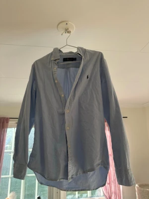 Ljusblå skjorta från Ralph Lauren - Klassisk ljusblå skjorta från Ralph Lauren med broderad logga på bröstet. Skjortan har lång ärm, knappar framtill och är tillverkad i bomull. Perfekt för en clean och stilren look.