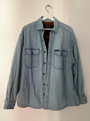 Ljusblå overshirt från Woolrich - Snygg ljusblå overshirt från Woolrich i denim-look. Två bröstfickor med knappar och klassisk krage. Insidan har ett mjukt foder i brunt och vinrött. Perfekt lager-på-lager-plagg för en avslappnad stil.