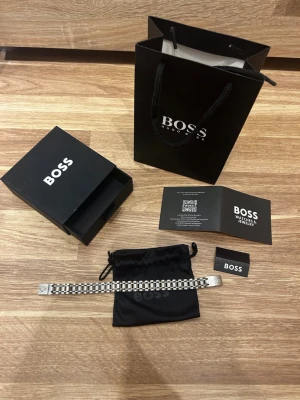 Silverfärgat armband från BOSS - Snyggt armband från BOSS i silverfärgad metall med ett stilrent, rutmönstrat band. Kommer med originalpåse, ask och presentpåse från BOSS. Perfekt accessoar för dig som gillar en modern och elegant look.