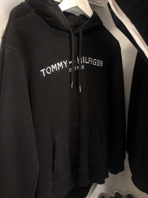 Svart hoodie från Tommy Hilfiger - Svart hoodie med huva och dragsnören från Tommy Hilfiger. Framtill finns en stor magficka och logotypen 'TOMMY HILFIGER EST. 1985' broderad i vitt, rött och blått. Perfekt för en avslappnad och stilren look.