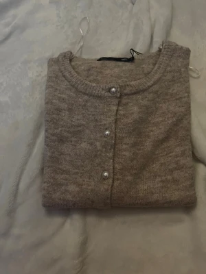 Beige kofta med pärlknappar - Mysig beige kofta med rund halsringning och dekorativa pärlknappar framtill. Koftan är stickad i mjukt material och har korta ärmar samt ribbade muddar vid ärmslut och nederkant. Perfekt för lager på lager och enkel att matcha. Koftan är i rosa beige färg.