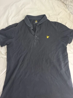 Svart pikétröja från Lyle & Scott - Klassisk svart pikétröja från Lyle & Scott med gul logga på bröstet. Tröjan har krage, knappar framtill och korta ärmar. Perfekt till jeans eller shorts för en clean och enkel stil.