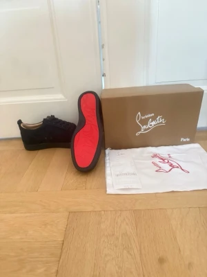 Svarta Louboutin sneakers med röd sula - Snygga svarta sneakers från Christian Louboutin i mocka med den ikoniska röda sulan. Skorna har lågt skaft, svarta skosnören och stilren design. Perfekta för dig som vill sticka ut med exklusiva detaljer och klassisk look.