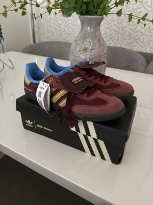 Adidas Wales Bonner burgundy sneakers - Snygga Adidas Wales Bonner sneakers i vinrött med gula detaljer och blå insida. Skorna har klassiska tre ränder på sidan, bred plös med Wales Bonner-logga och mörk sula. Materialet är en mix av mocka och textil. Perfekta för dig som vill sticka ut med stilrena detaljer.