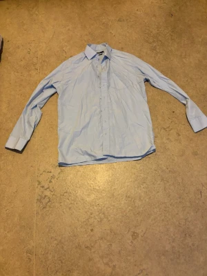 Ljusblå skjorta - Säljer en ljusblå skjorta med klassisk krage och långa ärmar. Skjortan har knäppning framtill och är perfekt för en clean och stilren look. Passar dig som gillar enkel och tidlös stil.