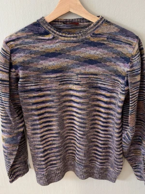 Missoni Melerad stickad tröja i lila toner - Säljer en stickad tröja med melerat mönster i lila, blått, beige och grått. Tröjan har rund halsringning, långa ärmar och ribbstickade muddar. Perfekt för dig som gillar unika färgkombinationer och en avslappnad stil. 0779