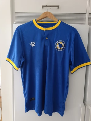 Bosnien & Hercegovina blå pikétröja (Kelme) – Präglat liljemönster - Säljer en pikétröja/matchtröja från Kelme i storlek M. Tröjan är i en riktigt fin blå färg med gula detaljer vid kragen och ärmarna, knäppning i halsen samt broderat förbundsmärke. Har dessutom ett riktigt snyggt ingraverat/präglat mönster med bosniska liljor (Ljiljan) över hela tyget.