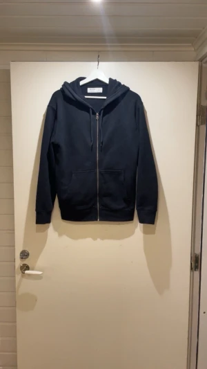 Svart zip-hoodie från Hollister XS - Svart hoodie från Hollister i relaxed fit med dragkedja framtill och två fickor. Klassisk huva med snörning och mjukt material som känns skönt mot huden. Perfekt för en chill och avslappnad stil.
