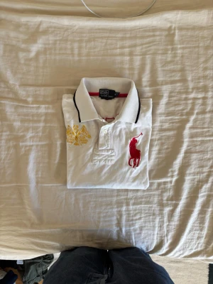Vit piké från Ralph lauren - Vit pikétröja med svart krage och röda detaljer. Stor röd broderad logga på bröstet och gul brodyr på andra sidan. Svarta och röda kontrastkanter på ärmar. Chief keif stil.