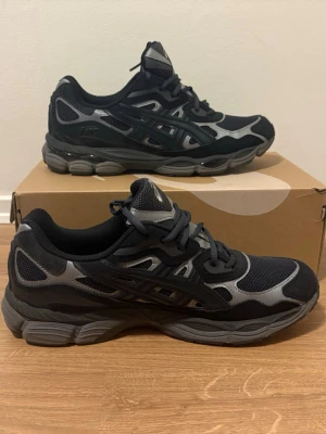 Svarta och grå Asics gel nyc - Snygga sneakers från Asics i svart och grått med mesh- och syntetpaneler. Skorna har en chunky sula och klassisk Asics-logga på sidan. Perfekta för dig som gillar sportig stil och vill ha bekväma skor till vardags.