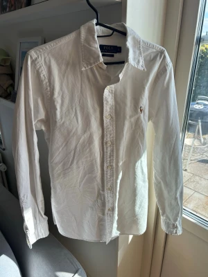 Vit skjorta Ralph Lauren Slim Fit - Klassisk vit skjorta från Polo Ralph Lauren i slim fit-modell. Skjortan har långa ärmar, knappar framtill och den ikoniska broderade loggan på bröstet. Tillverkad i mjuk bomull och passar perfekt till en clean och stilren look.